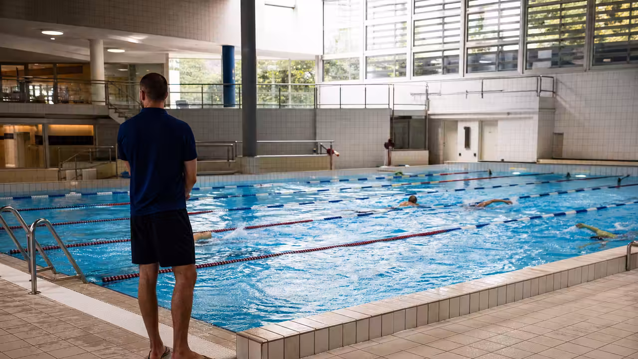 Lignes d’eau d’une piscine municipale calme, surveillées depuis le bord par un maître-nageur