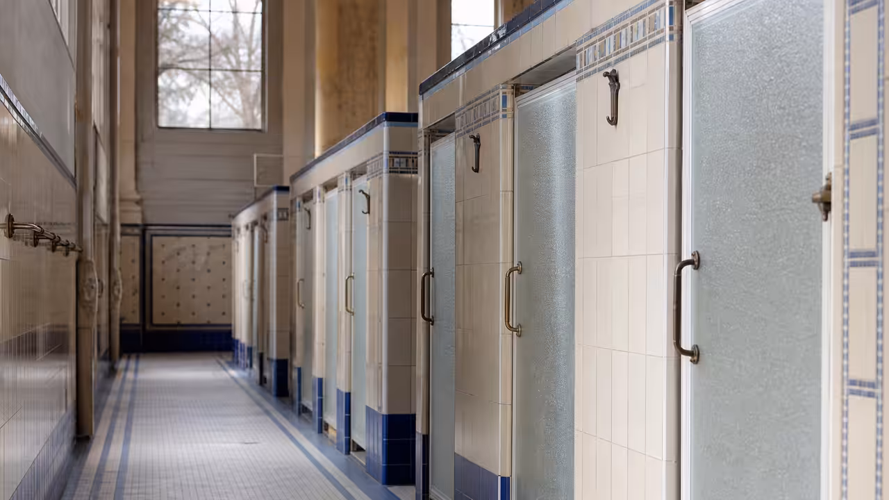 Cabines de bains-douches d’un équipement public ancien, avec carrelage clair et lumière naturelle.