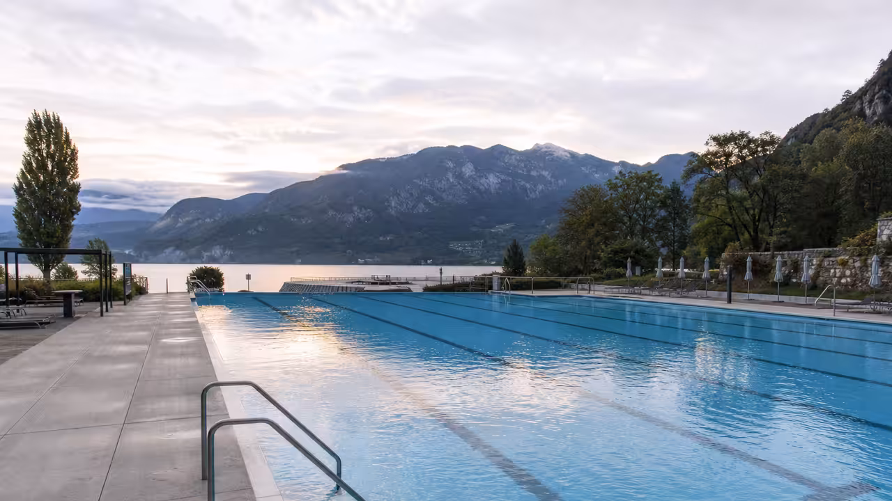 Bassin sportif extérieur vide avec lignes d’eau, gradins sobres et montagnes autour d’Annecy en arrière-plan