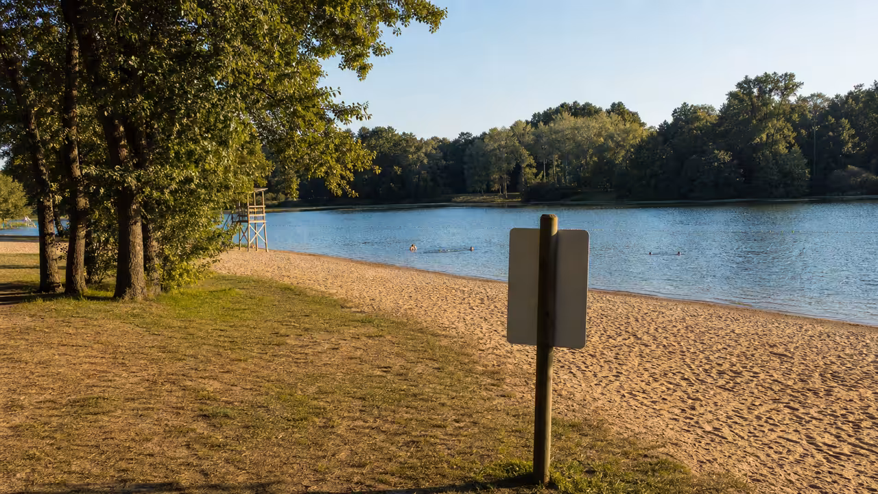 Zone de baignade aménagée au bord d’un plan d’eau, avec plage, panneaux de sécurité et baigneurs au loin