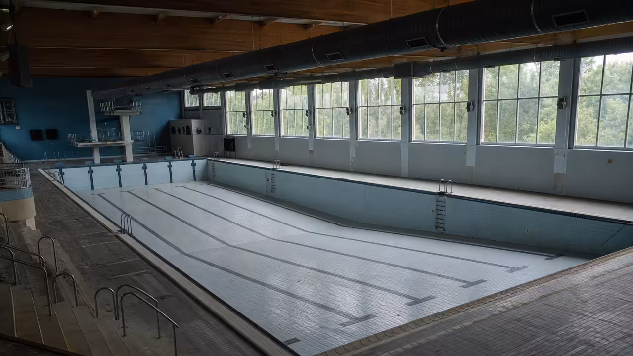 Bassin intérieur vide et carrelé d’un centre aquatique fermé, observé depuis les gradins sous lumière naturelle