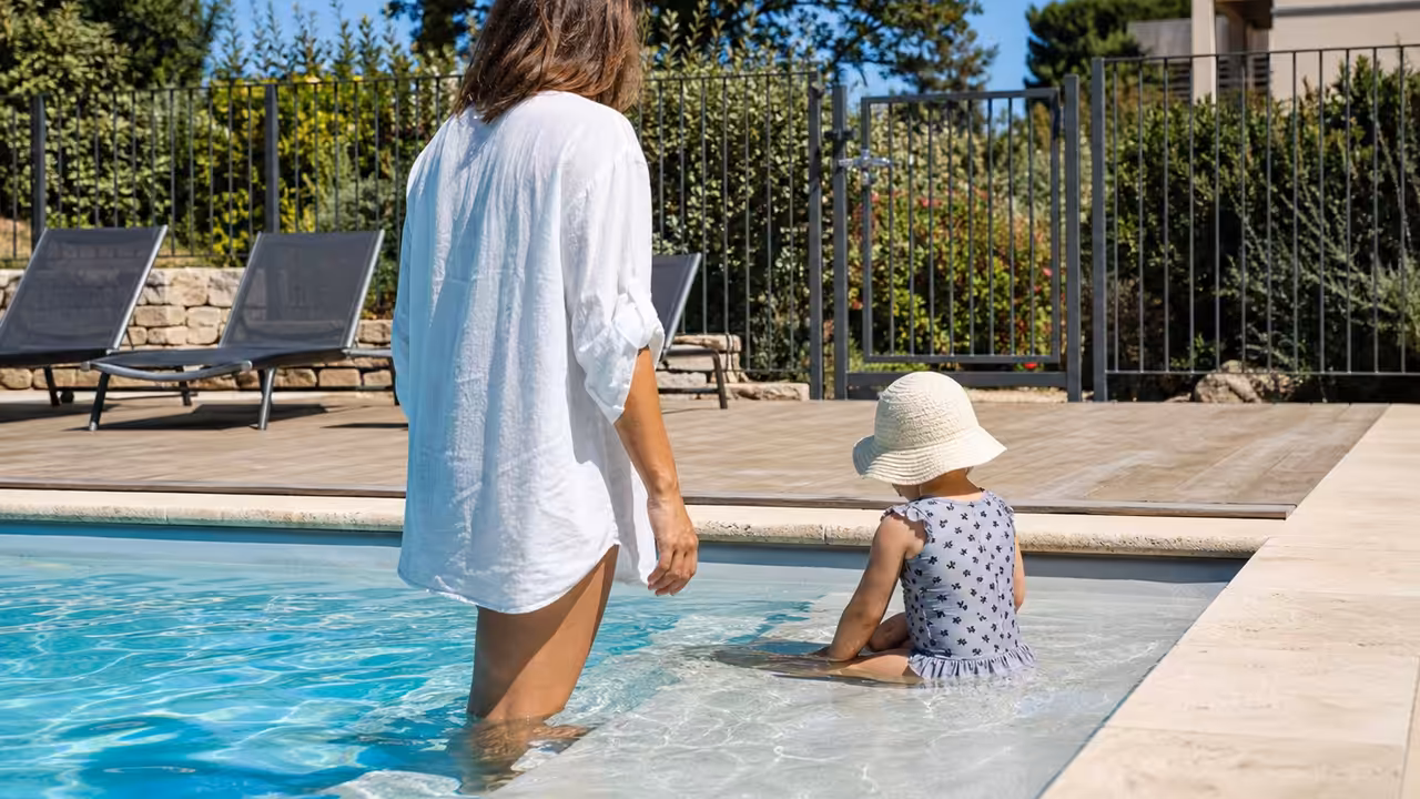 Un adulte surveille de près un jeune enfant jouant dans une piscine familiale sécurisée par une barrière.