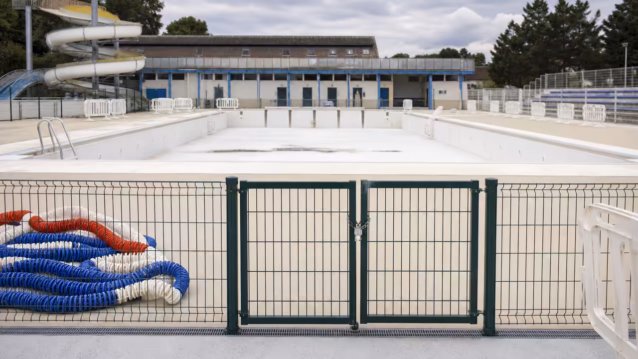 Portail fermé devant une piscine municipale extérieure vide, avec bassin et vestiaires en arrière-plan