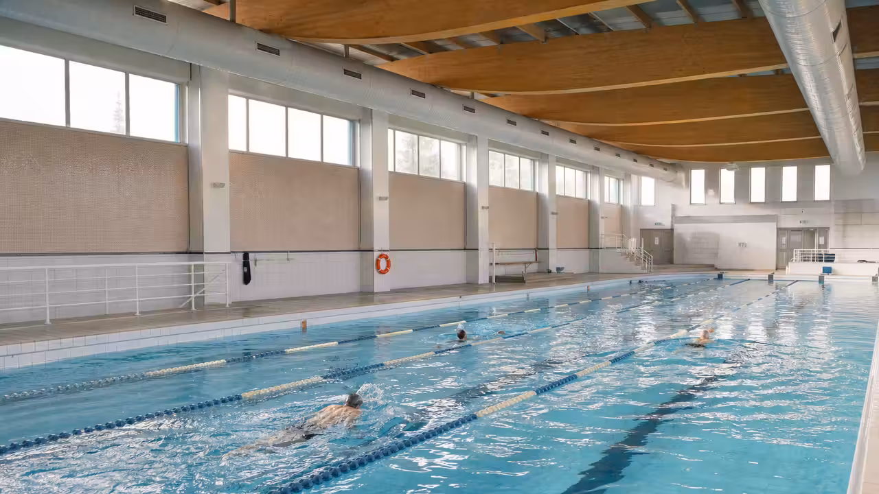 Bassin couvert d’une piscine municipale avec lignes d’eau, gradins sobres et nageurs vus de loin