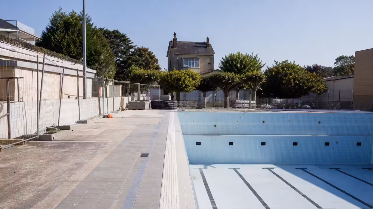 Bassin municipal vide en rénovation, avec plages carrelées et équipements techniques en arrière-plan