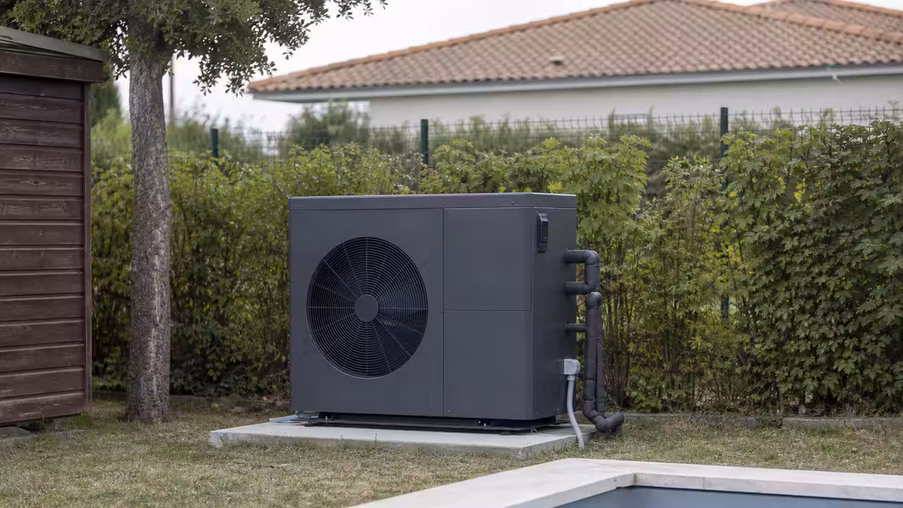 Pompe à chaleur de piscine sur une dalle béton au fond d'un jardin, ventilateur de face, haie et clôture en limite de terrain