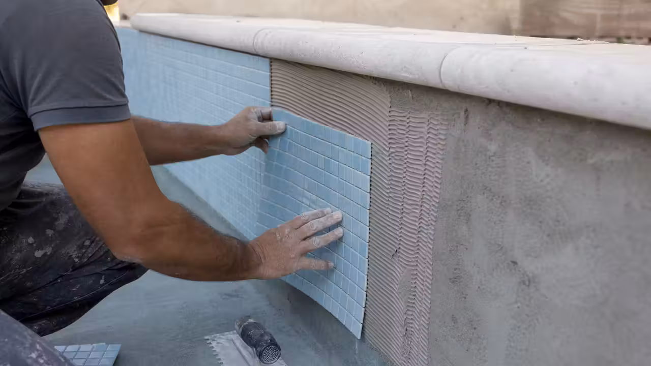 Poseur appliquant des mosaïques bleu clair sur la paroi d’un bassin maçonné vide avant jointoiement.