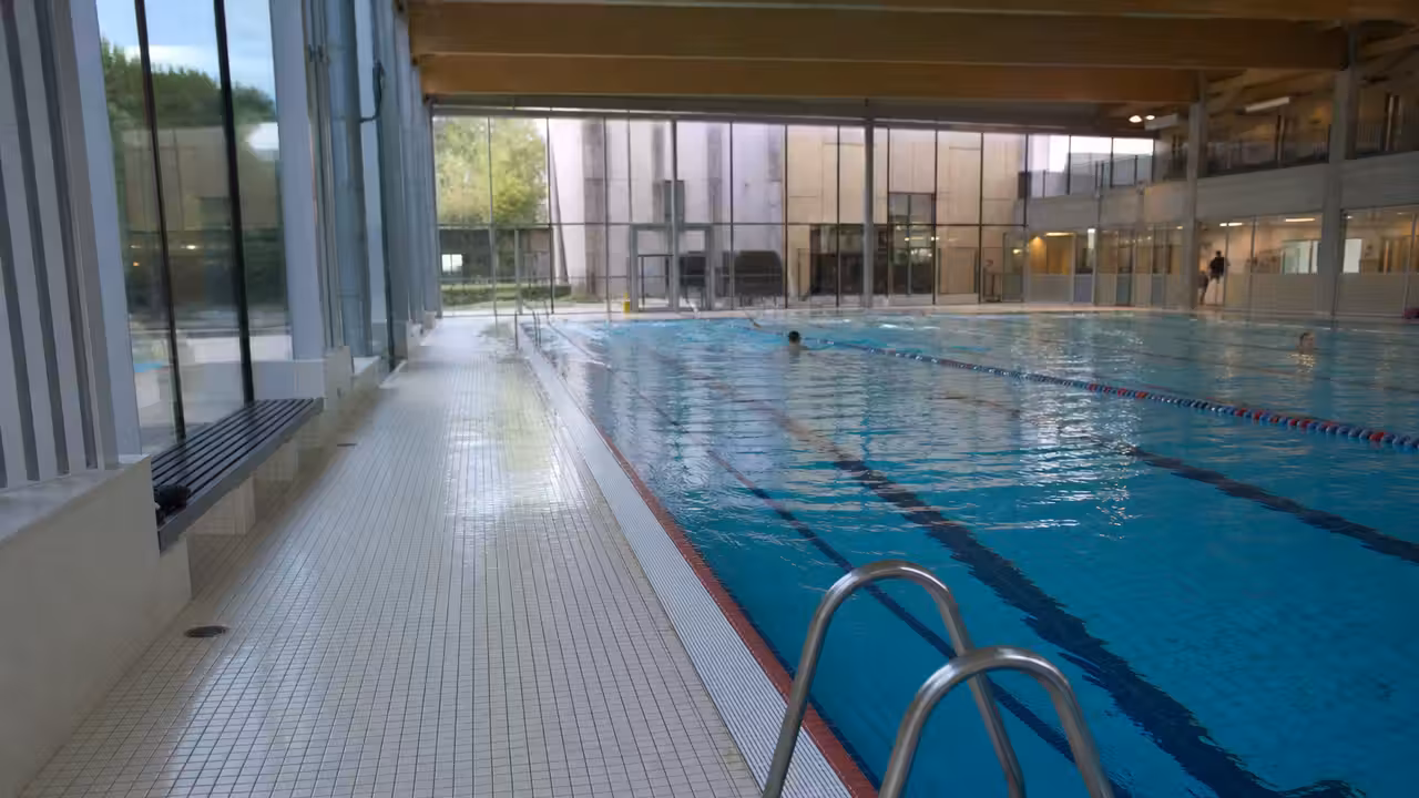 Bassin couvert municipal lumineux avec lignes d’eau, vestiaires vitrés et nageurs adultes au loin