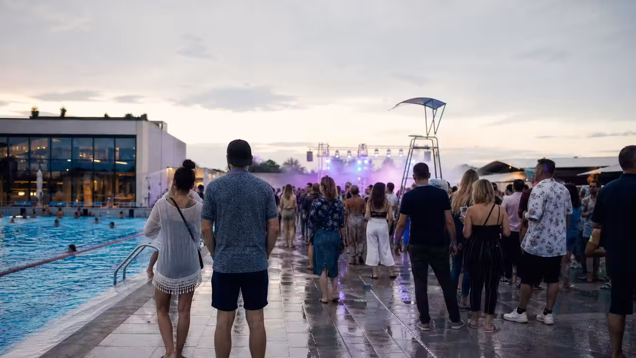 Scène de musique électronique au bord d’un bassin extérieur, avec public et éclairages en début de soirée