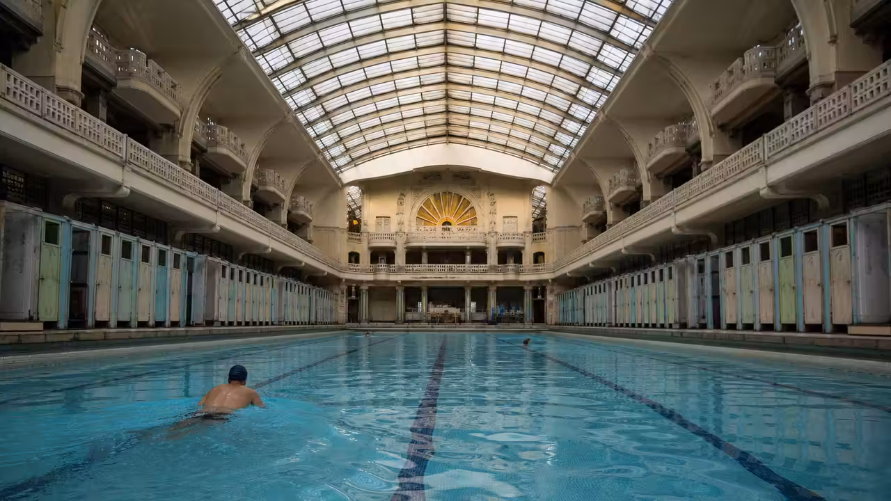 Bassin couvert Art déco avec verrière, cabines colorées et lignes d’eau dans une piscine parisienne