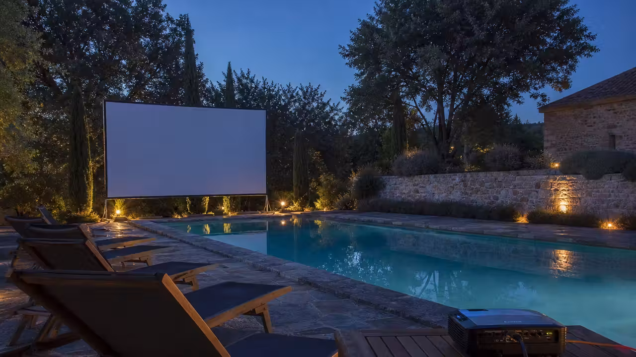 Écran de cinéma installé sur une terrasse de piscine éclairée, avec transats et bassin en arrière-plan