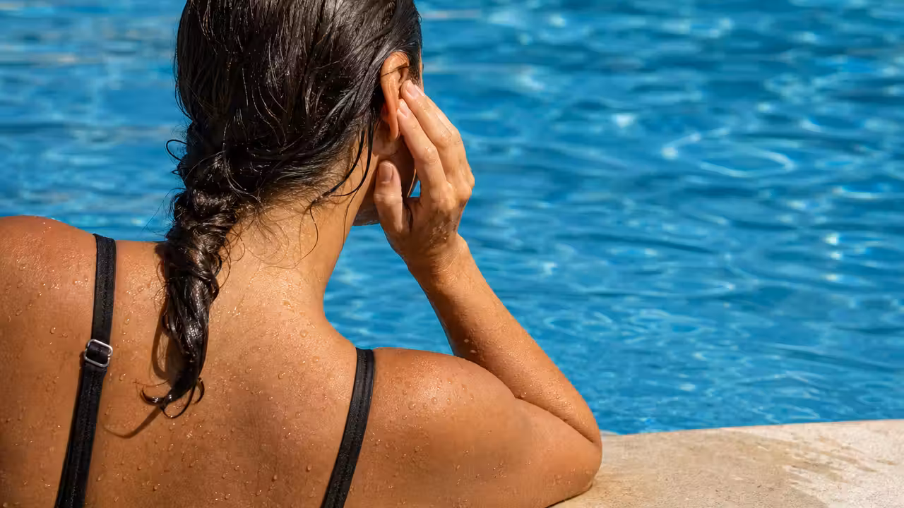 Enfant sortant la tête de l'eau d'une piscine, main portée près de l'oreille, lumière naturelle d'été