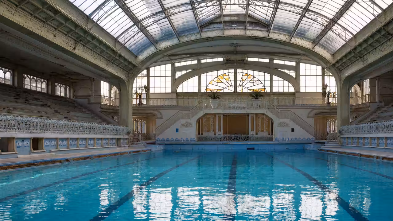 Grand bassin couvert de 50 mètres avec verrière, gradins vides et lignes d’eau dans une piscine publique patrimoniale