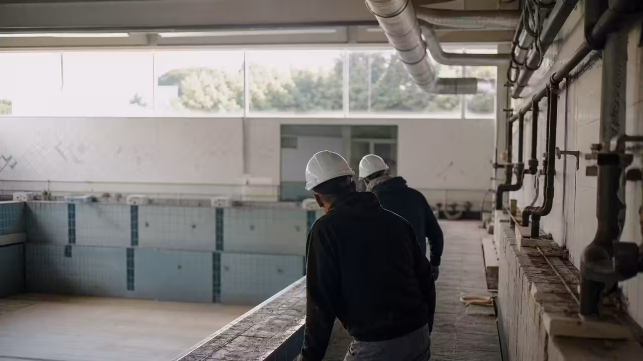 Bassin municipal vidé en rénovation, avec réseaux techniques et ventilation visibles dans un hall lumineux