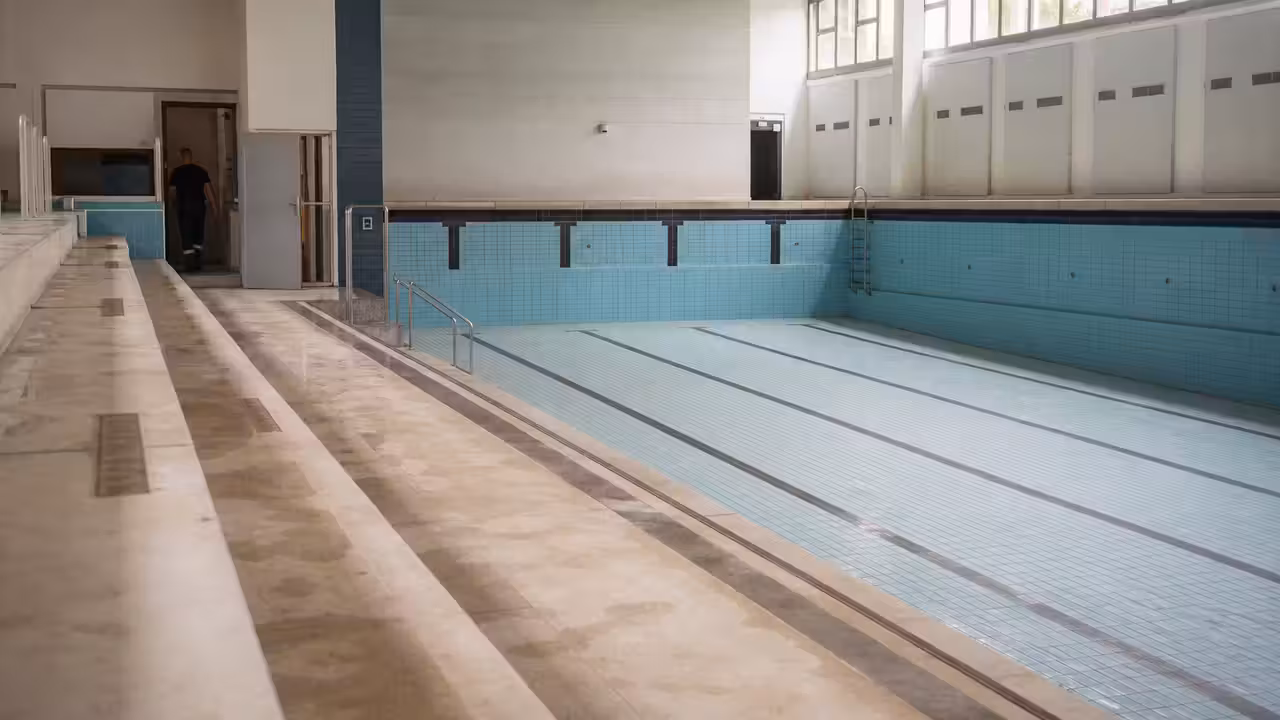 Bassin de piscine municipale vide avec lignes d’eau, gradins sobres et local technique aperçu en arrière-plan
