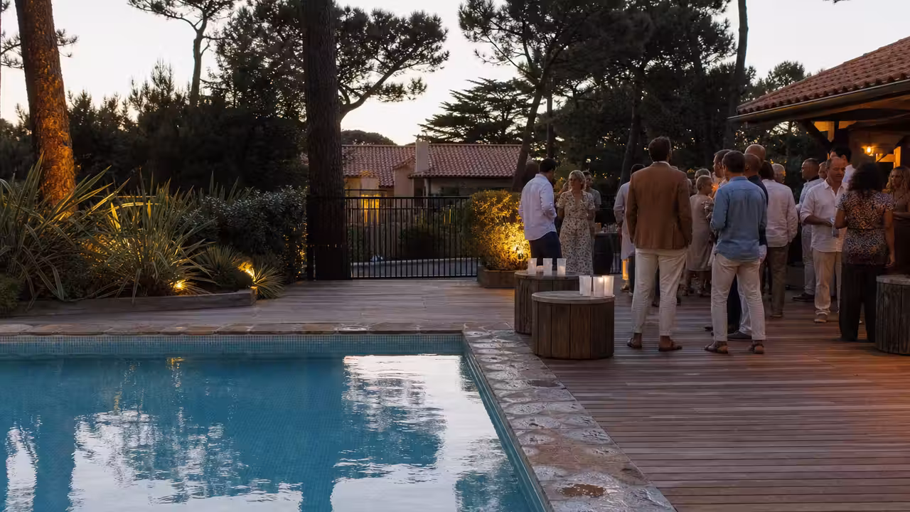 Piscine extérieure au crépuscule, terrasse éclairée et invités adultes réunis loin du bord de l’eau