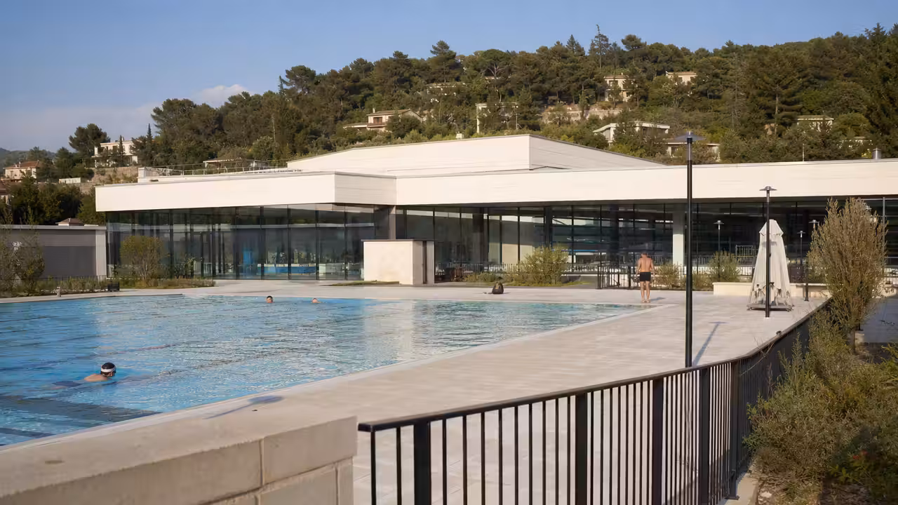 Vue en paysage d’un centre aquatique municipal moderne avec bassin extérieur, plages et collines en arrière-plan.