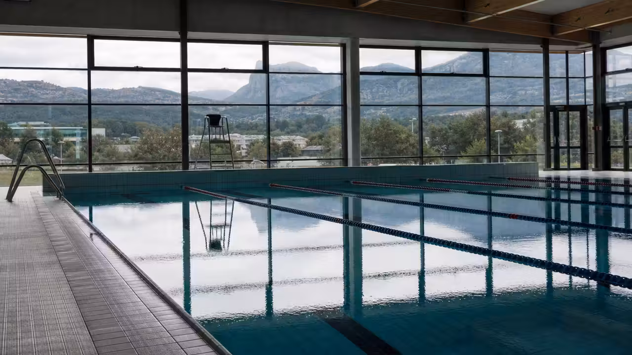 Bassin municipal couvert vide avec lignes d’eau, gradins sobres et reliefs alpins à l’arrière-plan