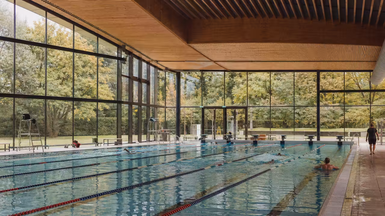 Bassin sportif couvert de 25 mètres avec lignes d’eau et larges baies vitrées dans un parc urbain