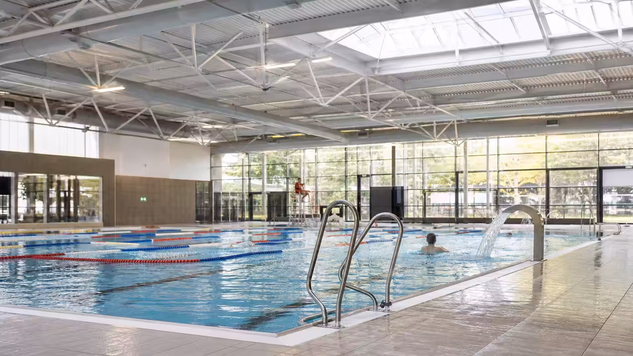 Bassin intérieur d’un centre aquatique public avec lignes d’eau, plafonds techniques et baigneurs au loin