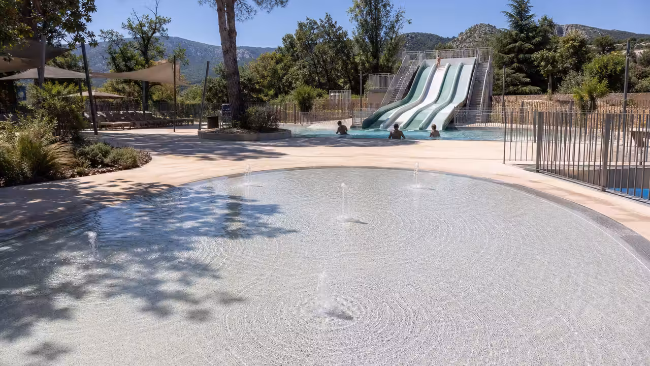 Espace extérieur d’une piscine publique avec jeux d’eau, plage minérale et toboggan aquatique