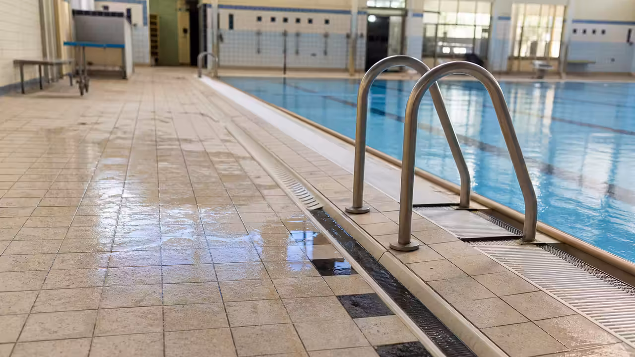Sol carrelé humide et vide autour d’un bassin intérieur, avec caniveaux et vestiaires en arrière-plan