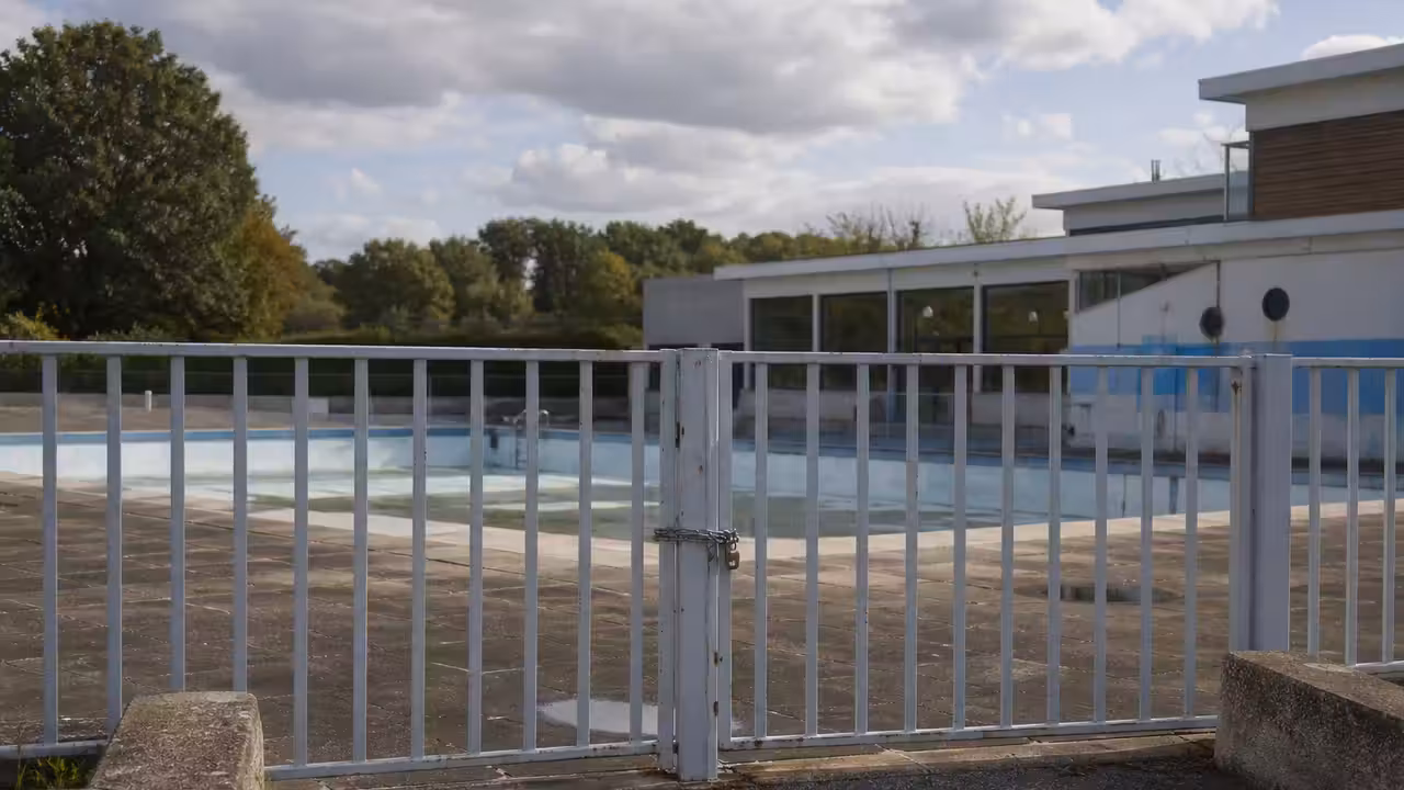 Entrée fermée d’une piscine municipale avec bassin extérieur vide et bâtiment technique en arrière-plan