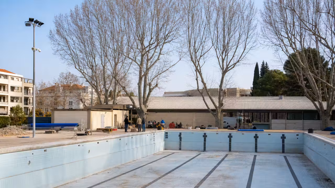 Bassin municipal vide en rénovation, avec plages carrelées et équipements techniques visibles à l'arrière-plan