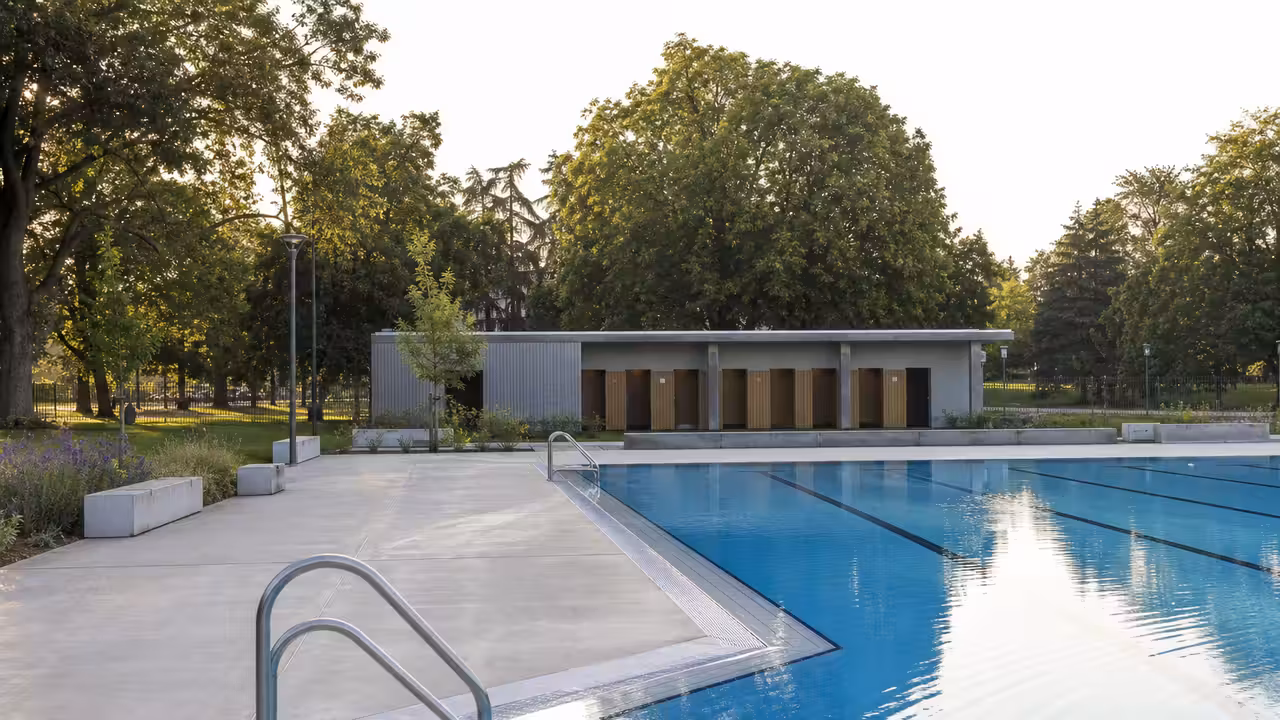 Bassin extérieur de piscine publique avec plages rénovées, locaux techniques discrets et parc arboré en arrière-plan