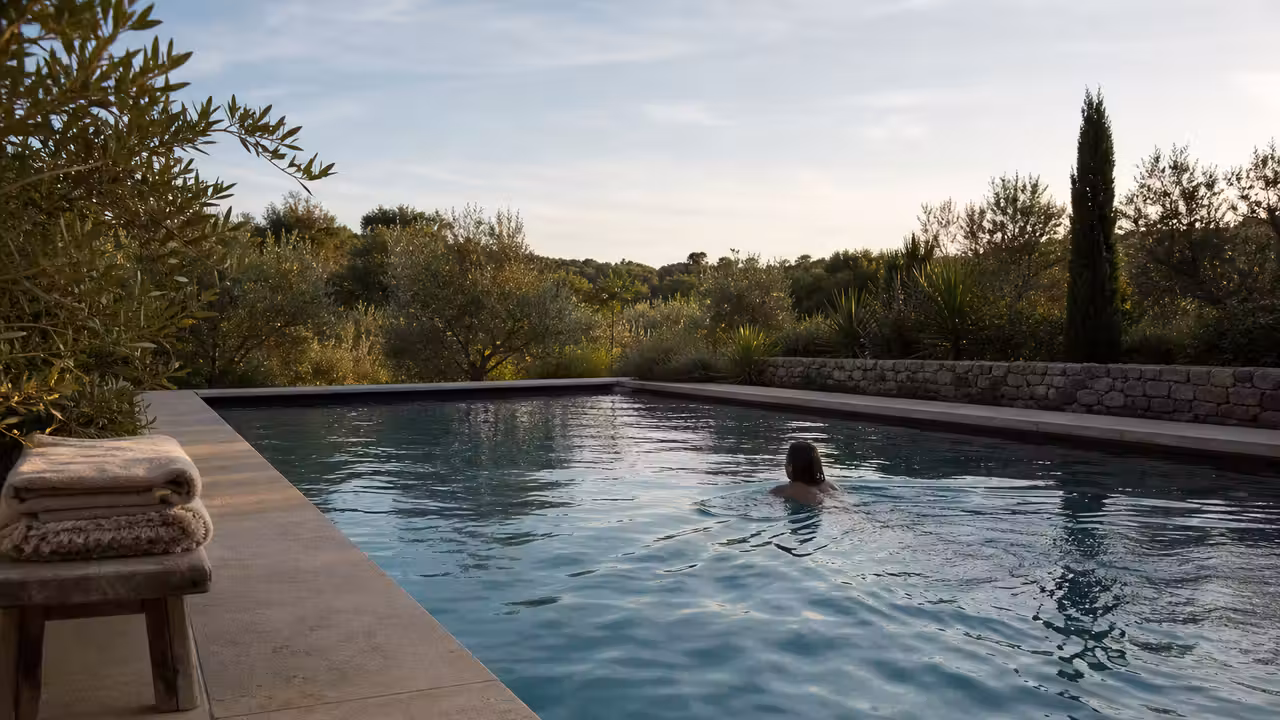 Adulte nageant lentement dans une piscine de jardin bordée d’une terrasse sobre, à la lumière du matin