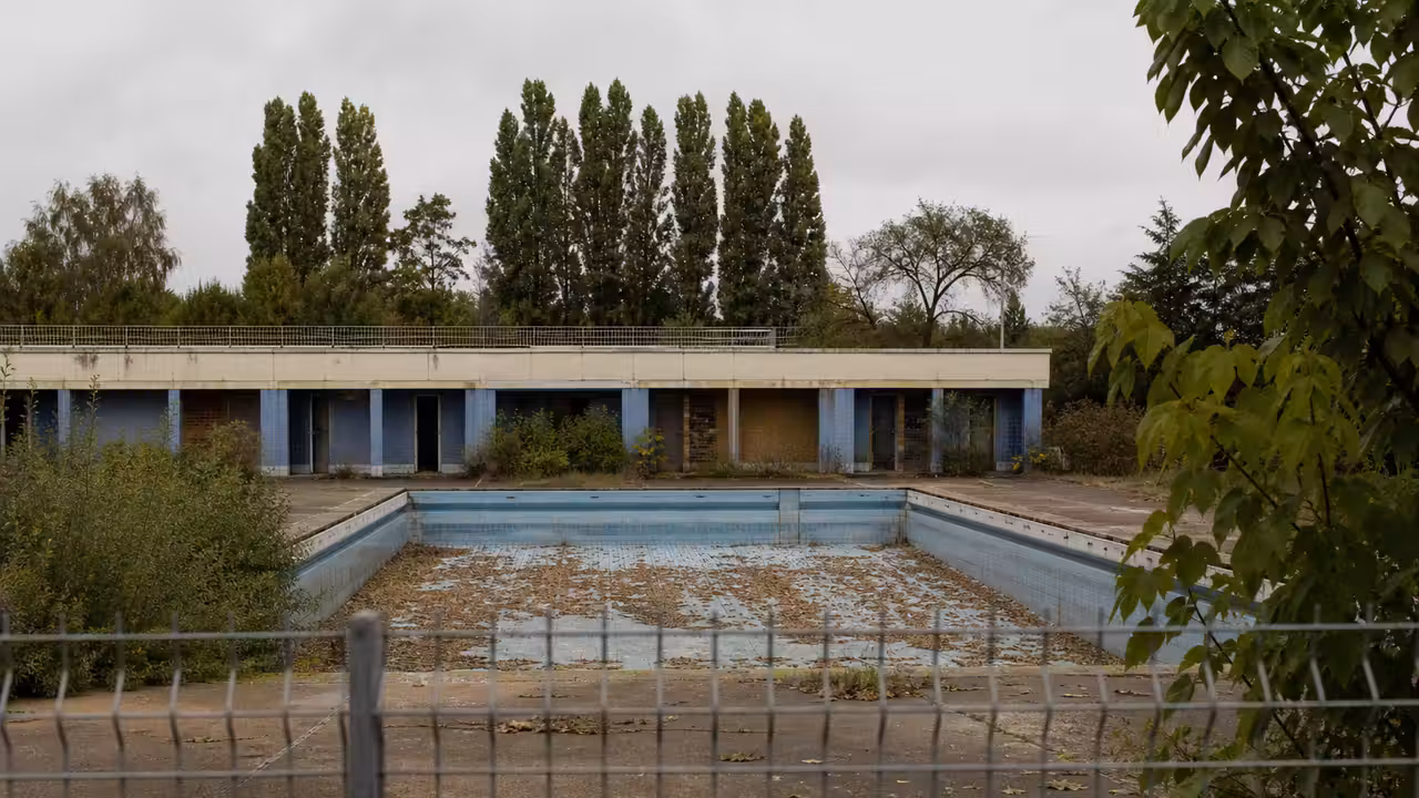 Ancienne piscine extérieure fermée, bassin vide et murs de béton marqués de graffitis, derrière une clôture