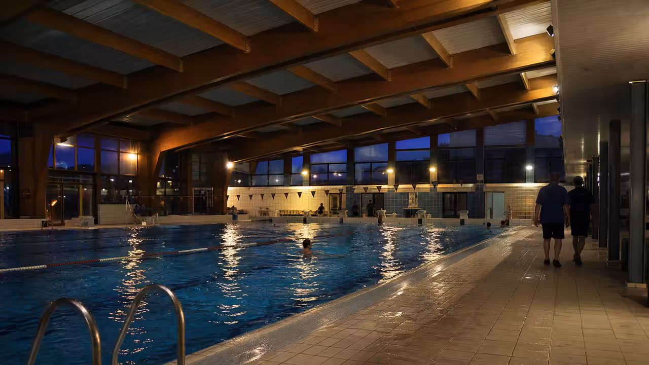 Bassin intérieur éclairé en soirée avec lignes d’eau, plages carrelées et silhouettes de nageurs au loin