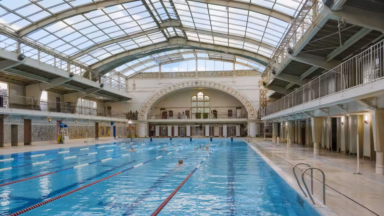 Lignes d’eau d’un bassin municipal de 50 mètres sous un toit vitré, avec nageurs au loin