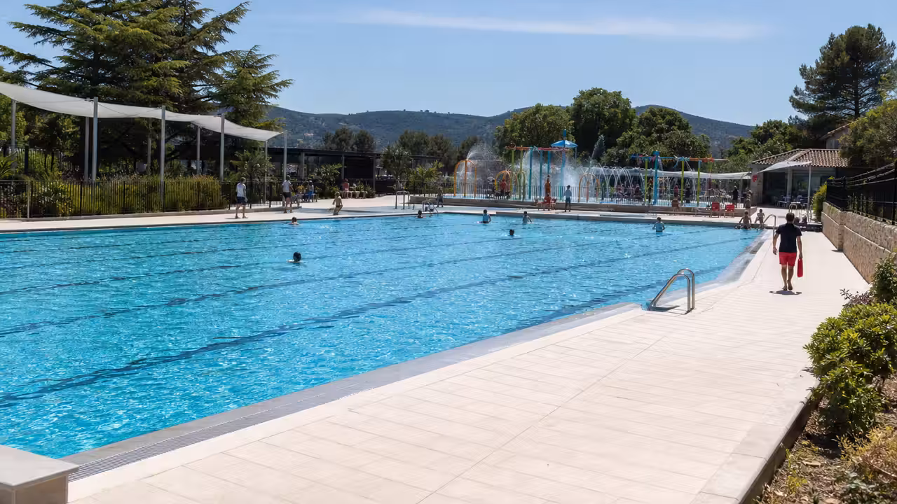 Le bassin extérieur rénové d’une piscine municipale avec plages claires et espace de jeux d’eau en arrière-plan
