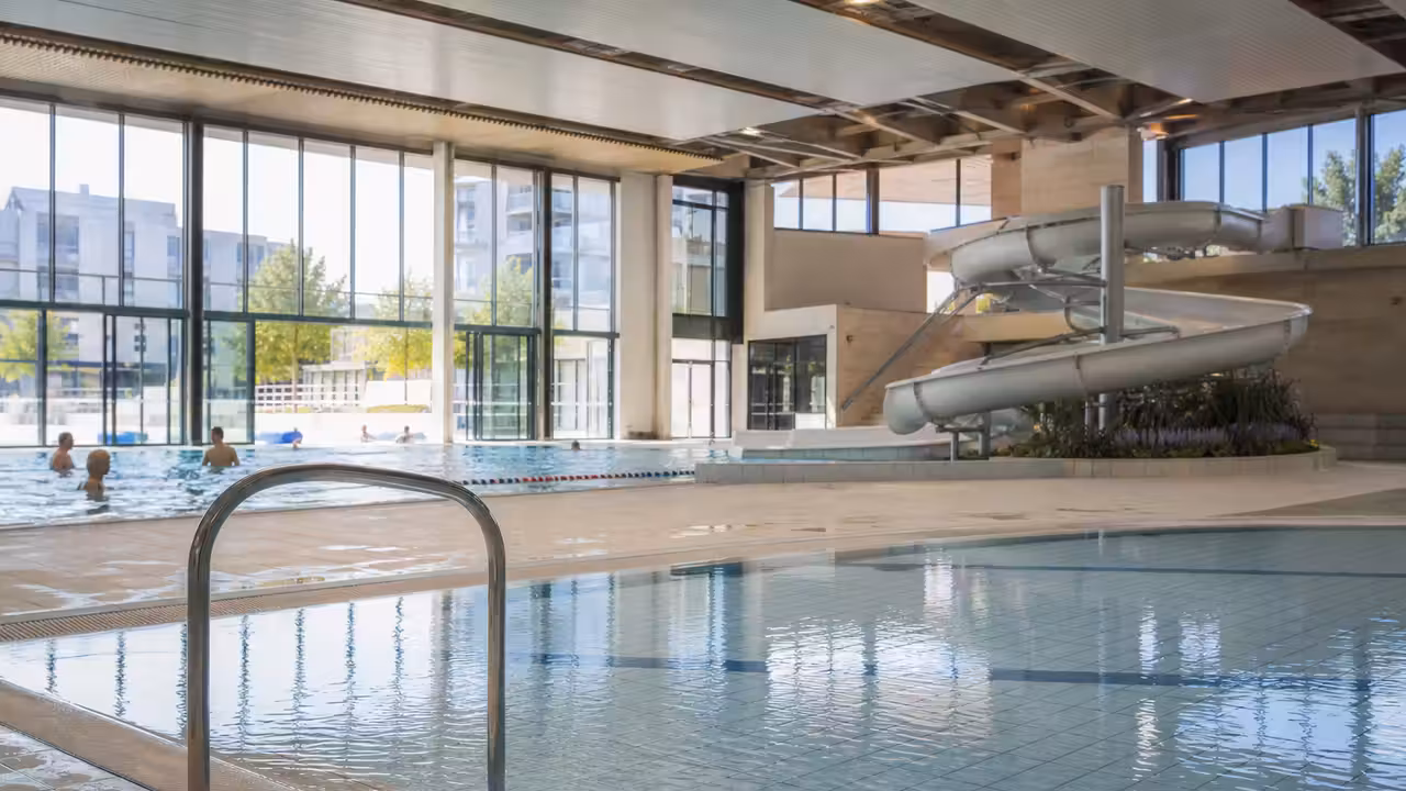 Les deux espaces de baignade rénovés d’un centre aquatique municipal, éclairés par de grandes baies vitrées.