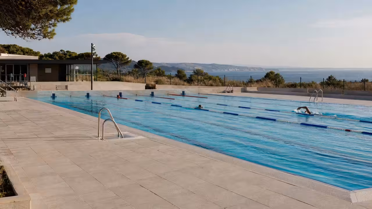 Bassin municipal extérieur bordé de plages minérales, avec lignes d’eau et locaux techniques en arrière-plan
