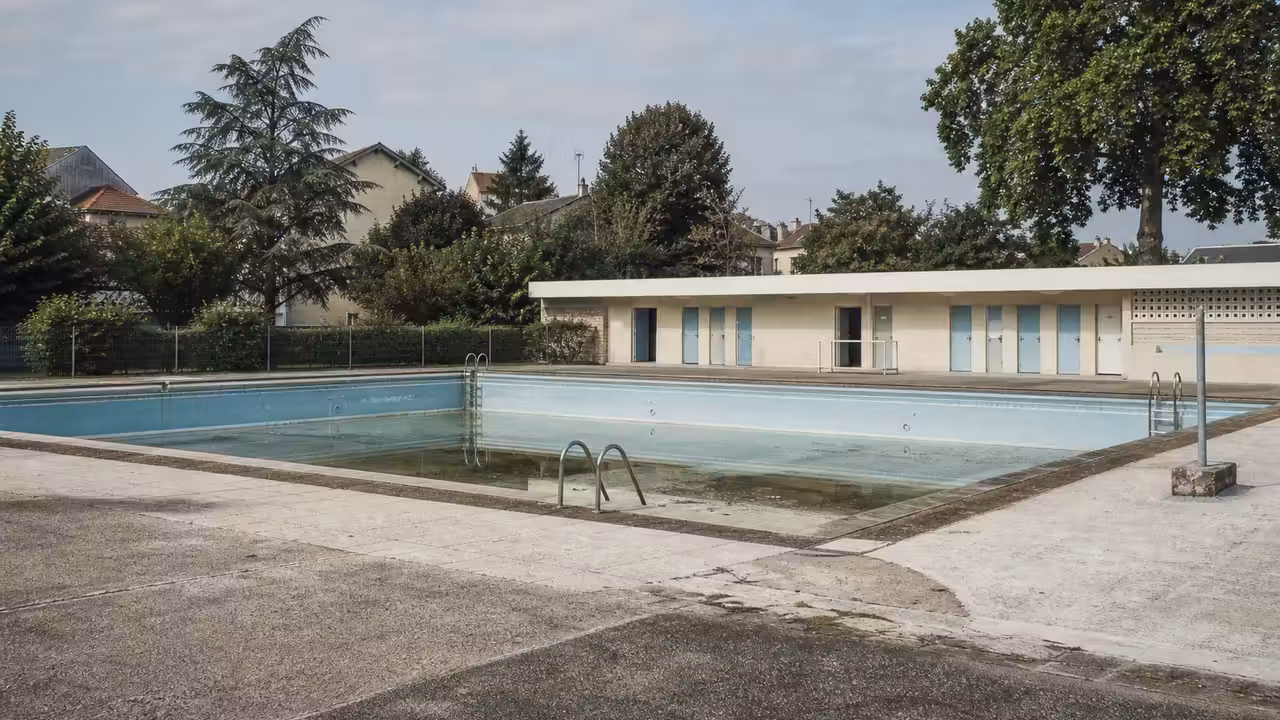 Ancien bassin extérieur municipal vide, entouré de plages minérales et de vestiaires à rénover