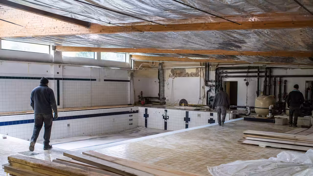 Halle de piscine publique en rénovation, avec bassins vides, charpente apparente et équipements techniques