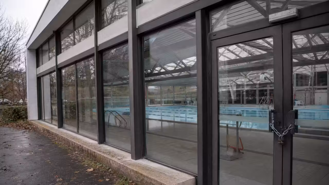 Entrée fermée d’une piscine municipale, avec un bassin intérieur vide visible derrière les baies vitrées