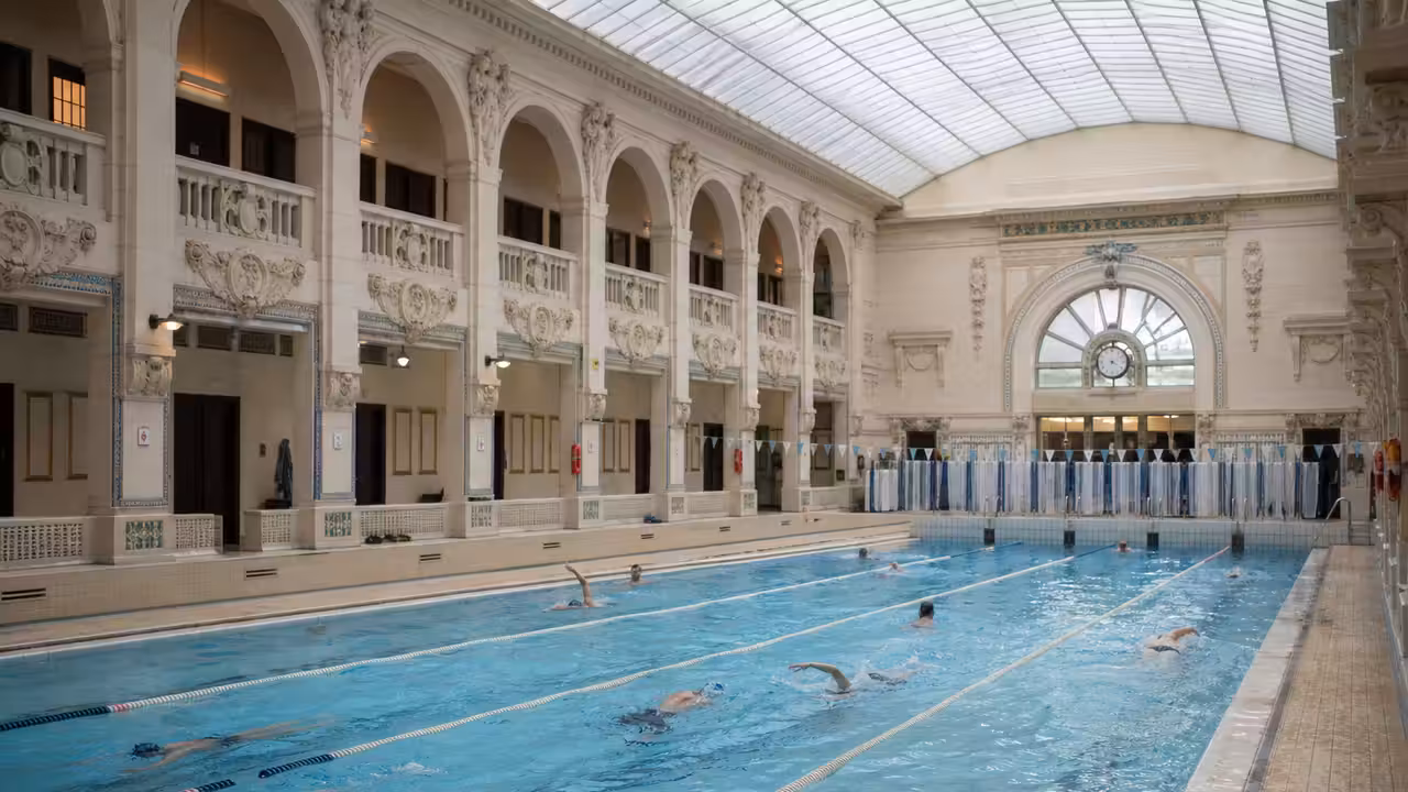 Nageurs adultes dans les lignes d’un bassin couvert parisien éclairé par une verrière historique