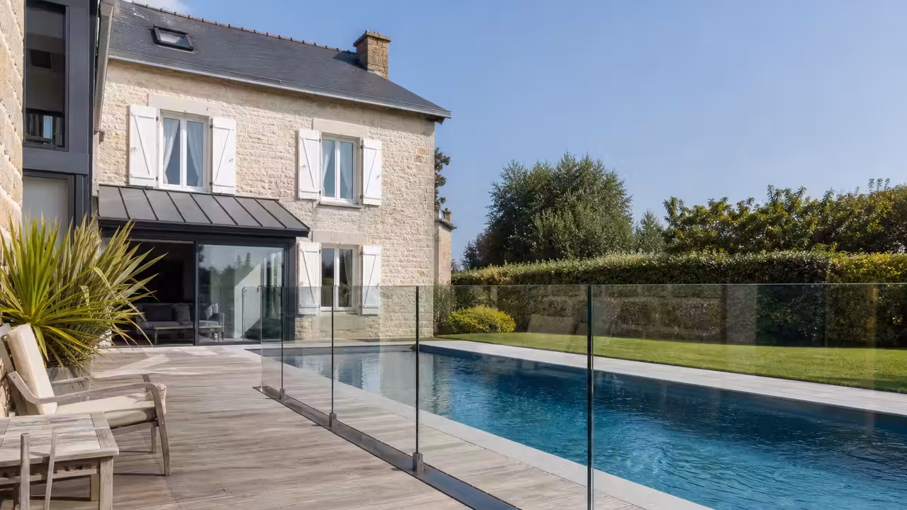 Piscine enterrée longeant la terrasse en bois d'une maison en pierre, barrière de protection en verre entre les deux