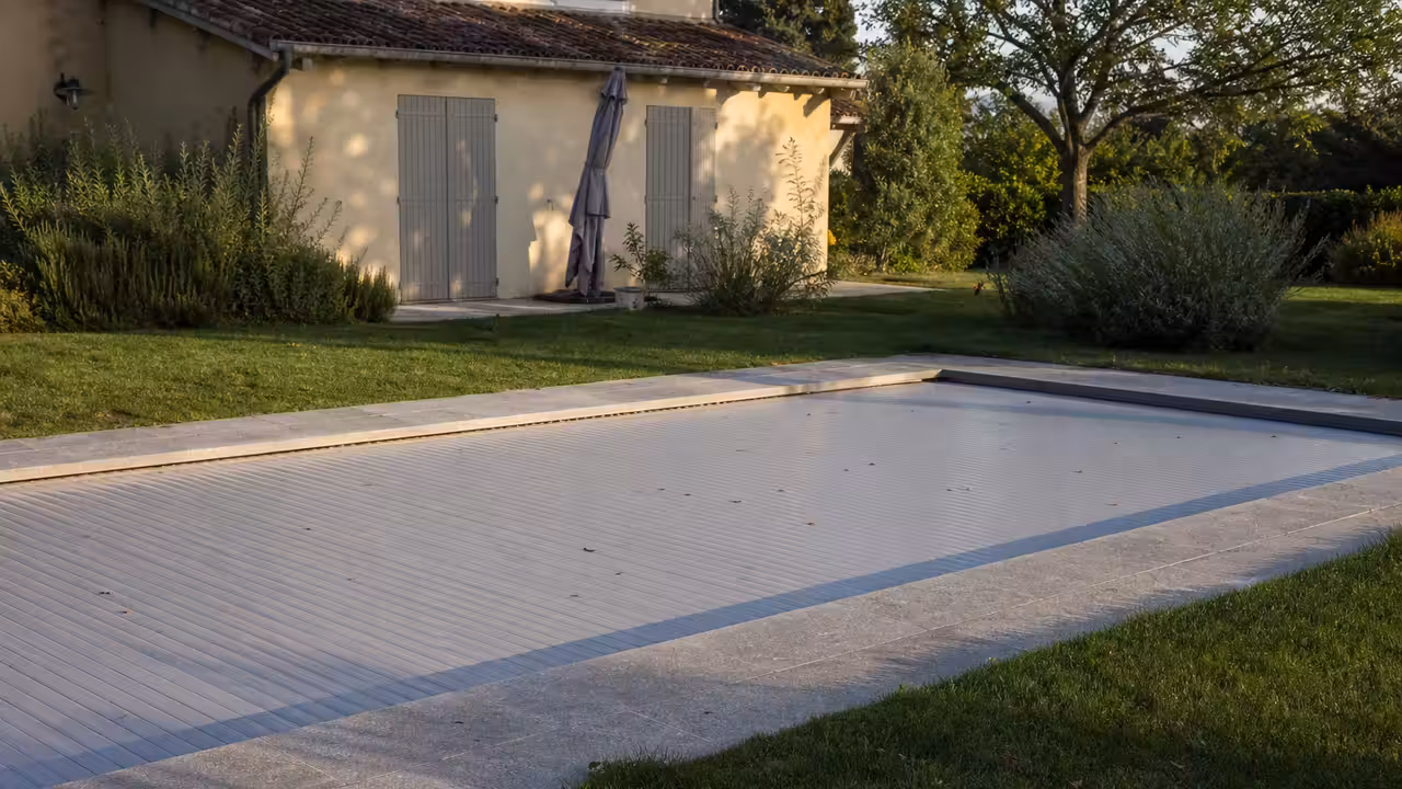 Piscine enterrée entièrement fermée par un volet à lattes dans un jardin désert, volets de la maison clos en fin de journée