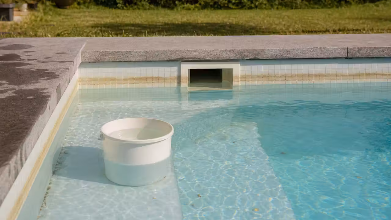Seau blanc posé sur la marche immergée d’une piscine privée, niveau de l’eau nettement descendu sous la bouche du skimmer