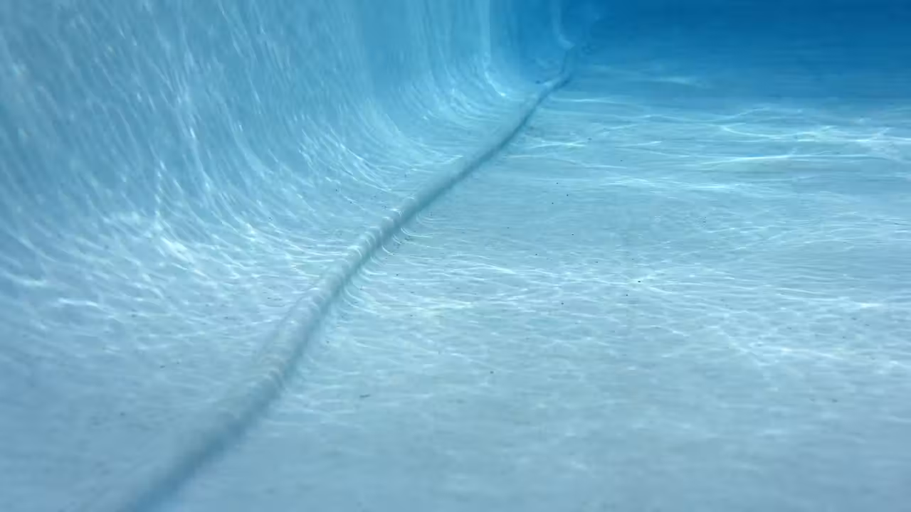 Fond d'une piscine en liner bleu clair vu à travers l'eau, avec un pli marqué courant le long du fond près de la paroi