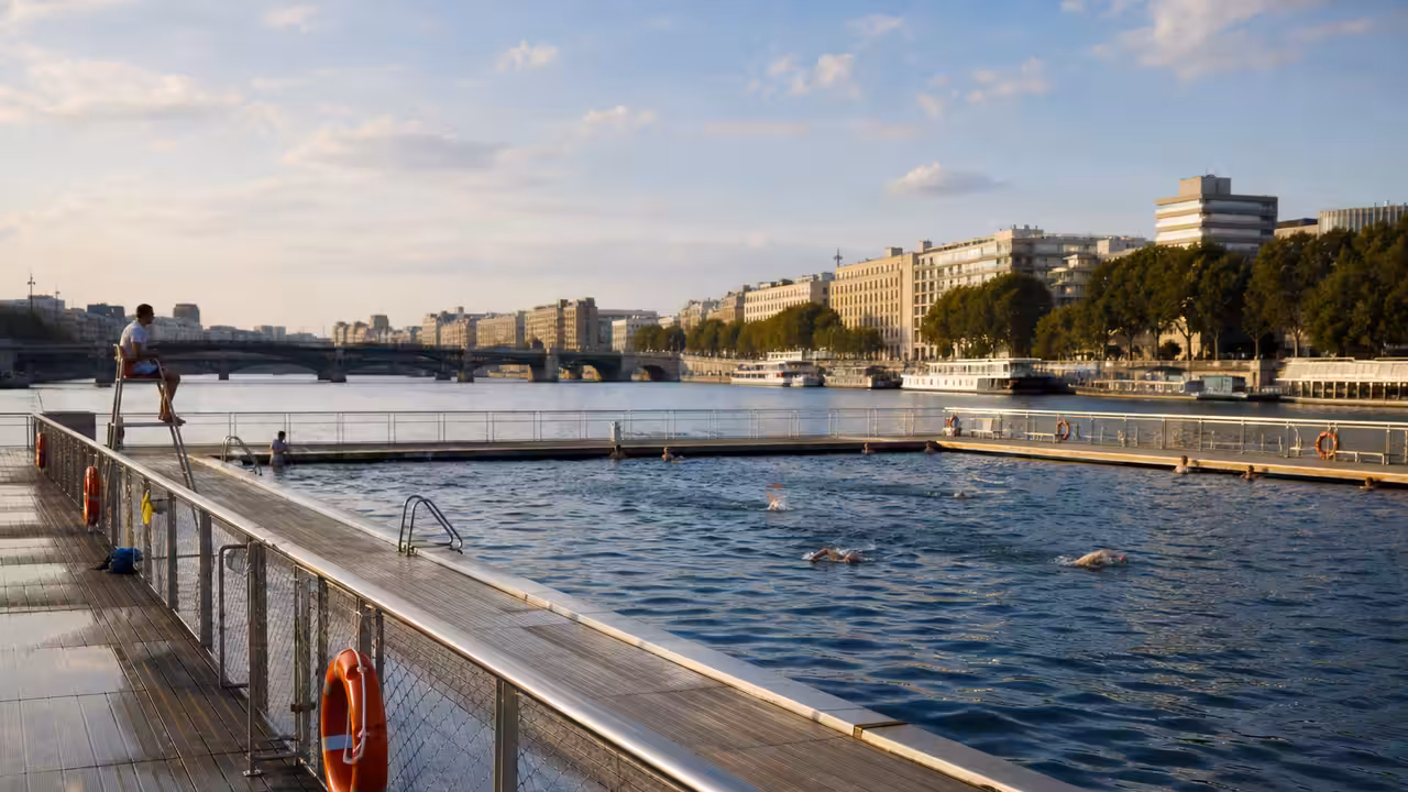Bassin public flottant sur un plan d’eau urbain, avec terrasse, garde-corps et skyline en arrière-plan