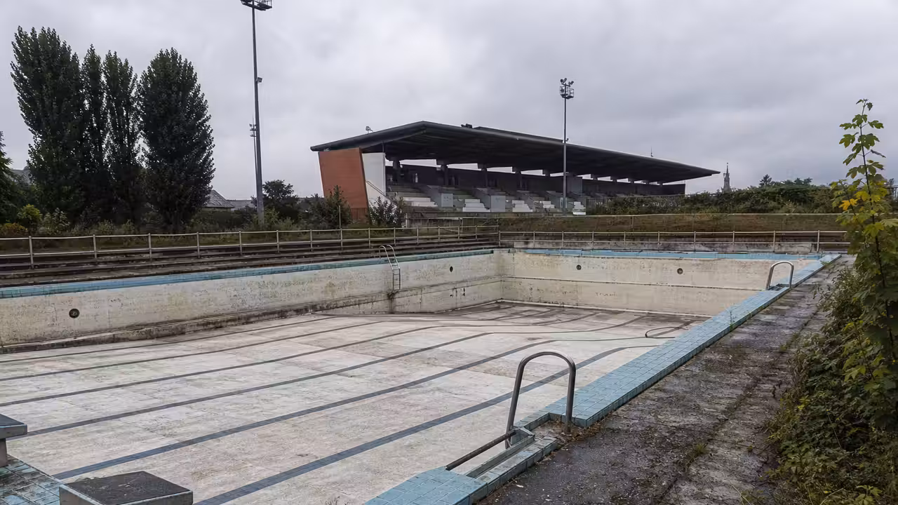 Bassin municipal extérieur vide, gradins anciens et végétation discrète autour d’un équipement fermé