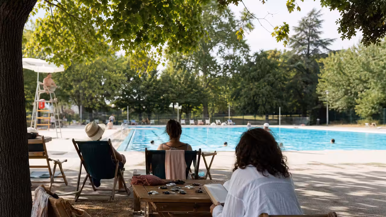 Espace extérieur d’une piscine municipale avec chaises longues, jeux de société et livres sur des tables ombragées