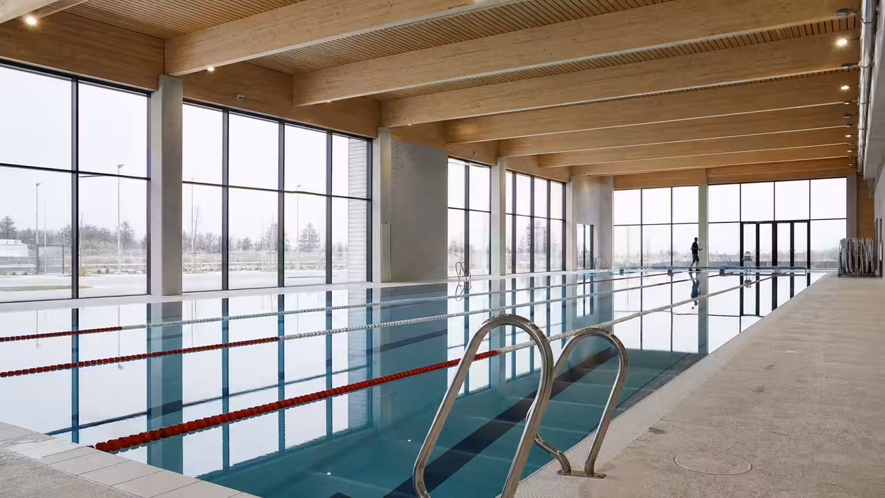 Bassin sportif couvert vide avec lignes d’eau, grandes baies vitrées et gradins d’une piscine municipale