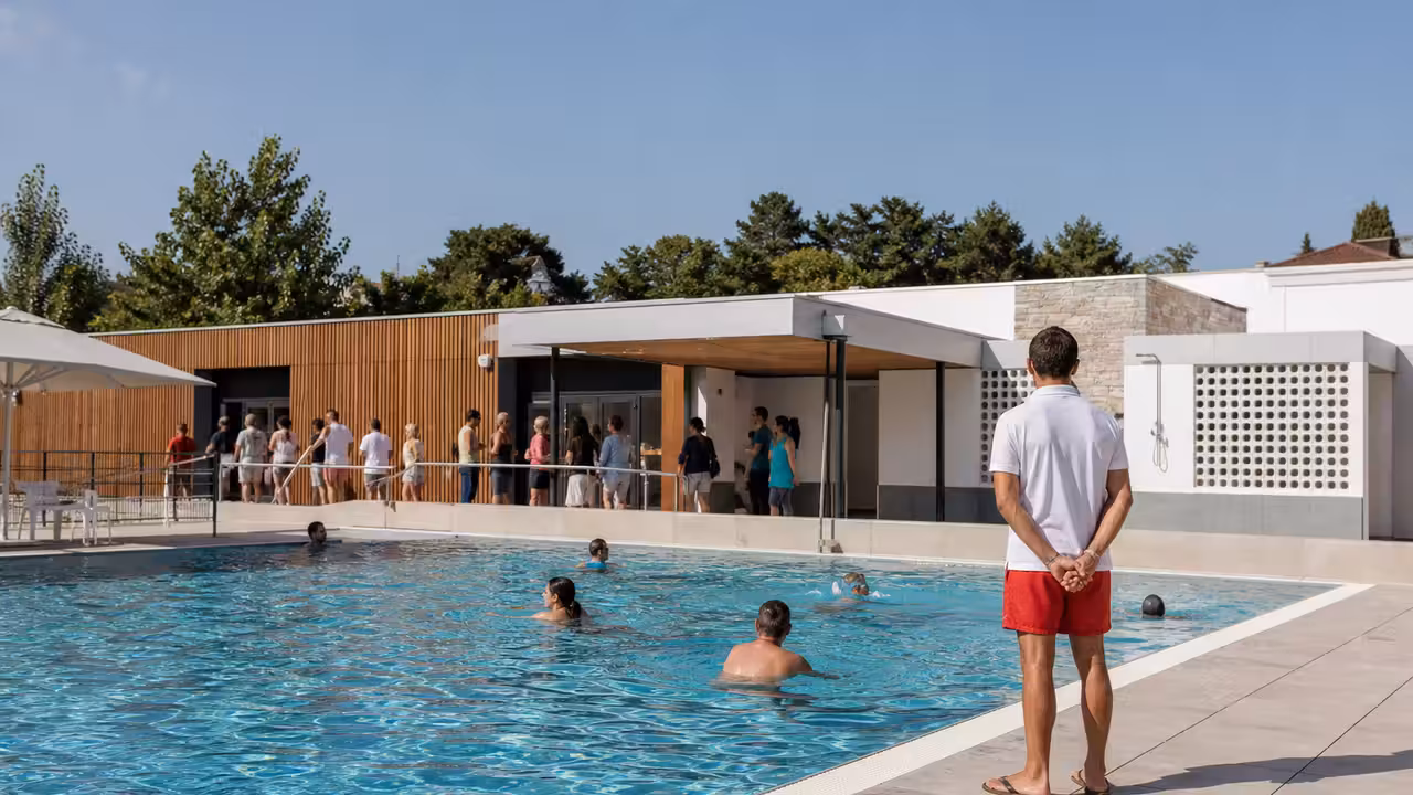 Bassin extérieur d’une piscine municipale en été, avec nageurs et maître-nageur au bord de l’eau