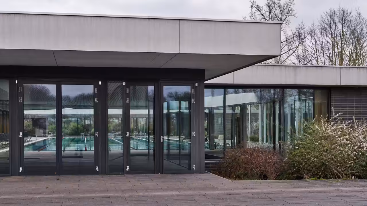 Entrée vitrée d’une piscine municipale fermée temporairement, avec bassin aperçu à travers les baies